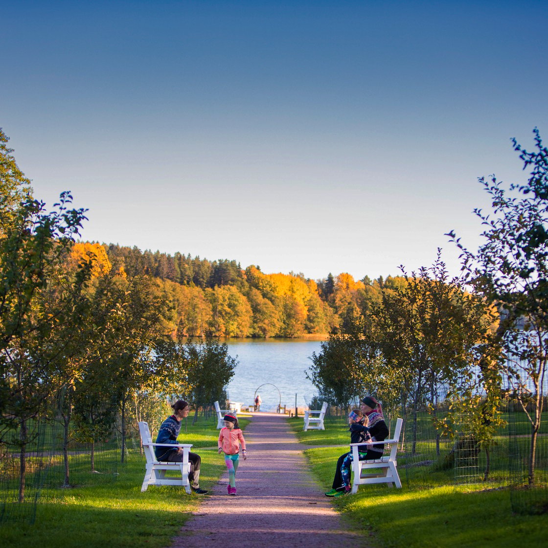 Kuva Wohls Gårdin rannalta. Kuvassa hiekkatie rannalle, jonka varrella muutamia valkoisia penkkejä. Kahdella penkillä istuu ihmisiä, hiekkatiellä kävelee lapsi.