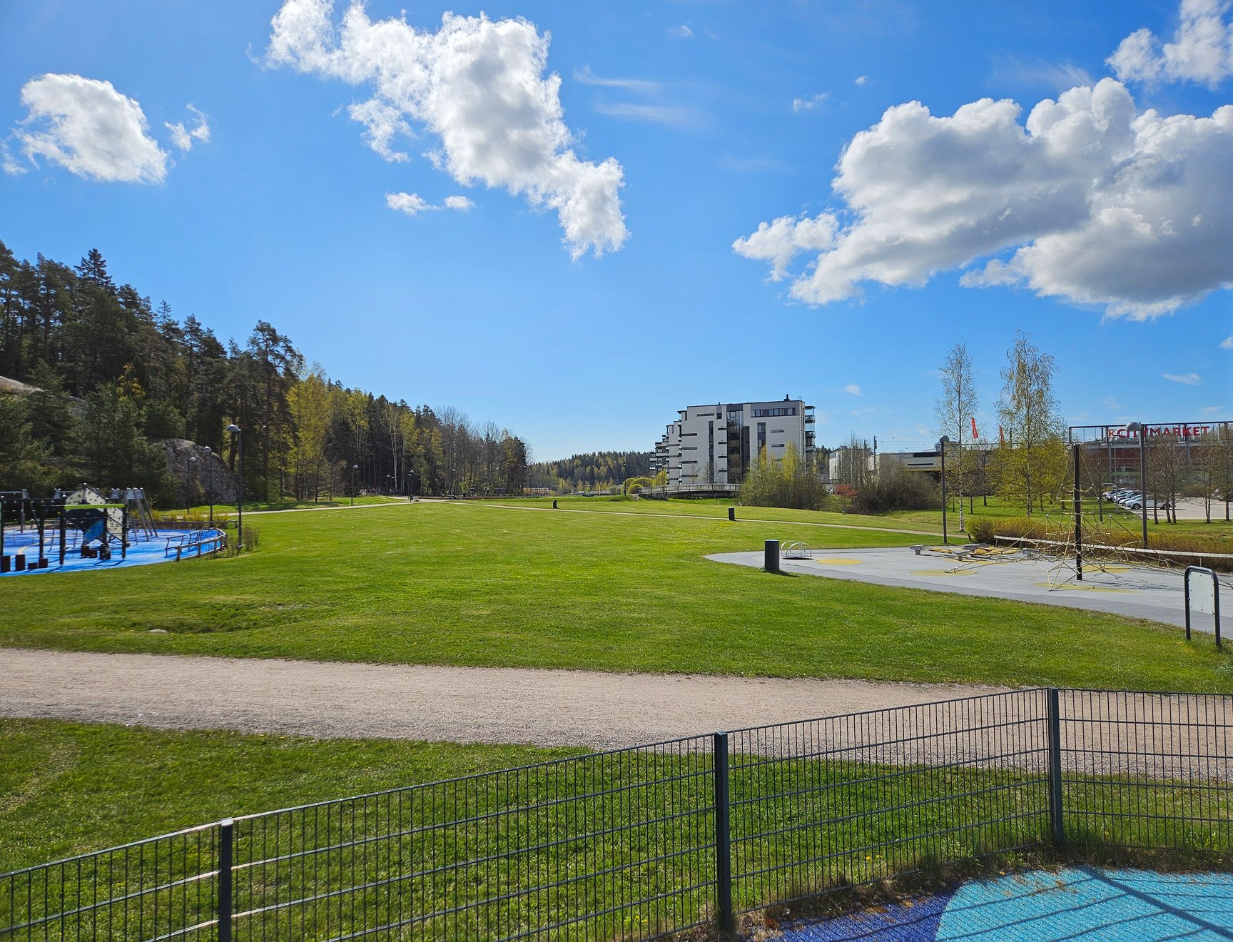 Kirkkonummen Kirsikkapuisto aurinkoisena kesäpäivänä. Näköpiirissä pääasiassa nurmialuetta, taustalla kerrostaloja ja edustalla hieman leikkipuistoja