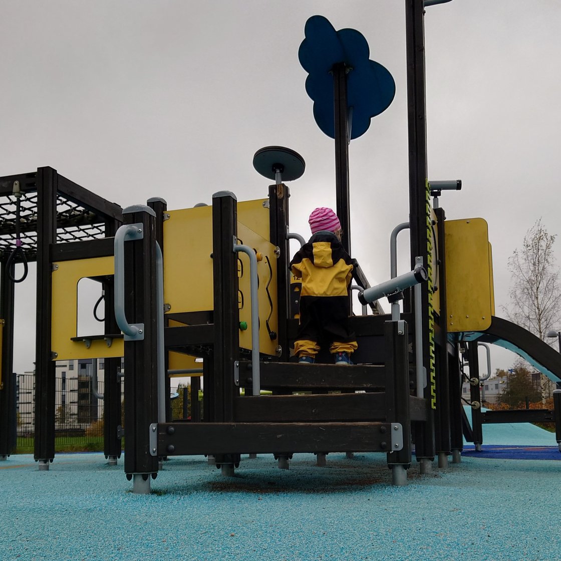 Pieni lapsi haalari päällä kiipeämässä leikkitelineeseen, jossa on liukumäki ja erinäisiä kiipeilytelineitä