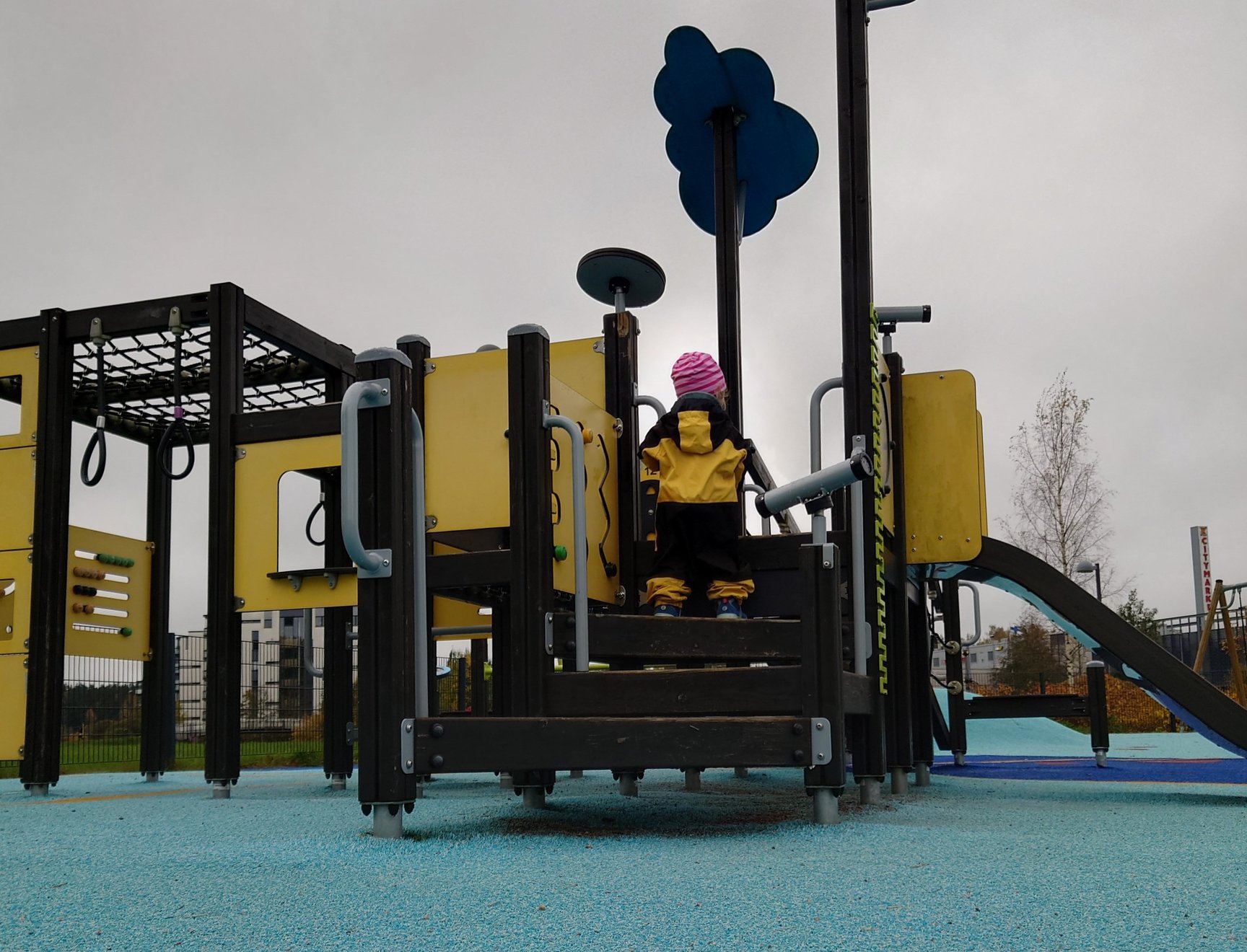 Pieni lapsi haalari päällä kiipeämässä leikkitelineeseen, jossa on liukumäki ja erinäisiä kiipeilytelineitä