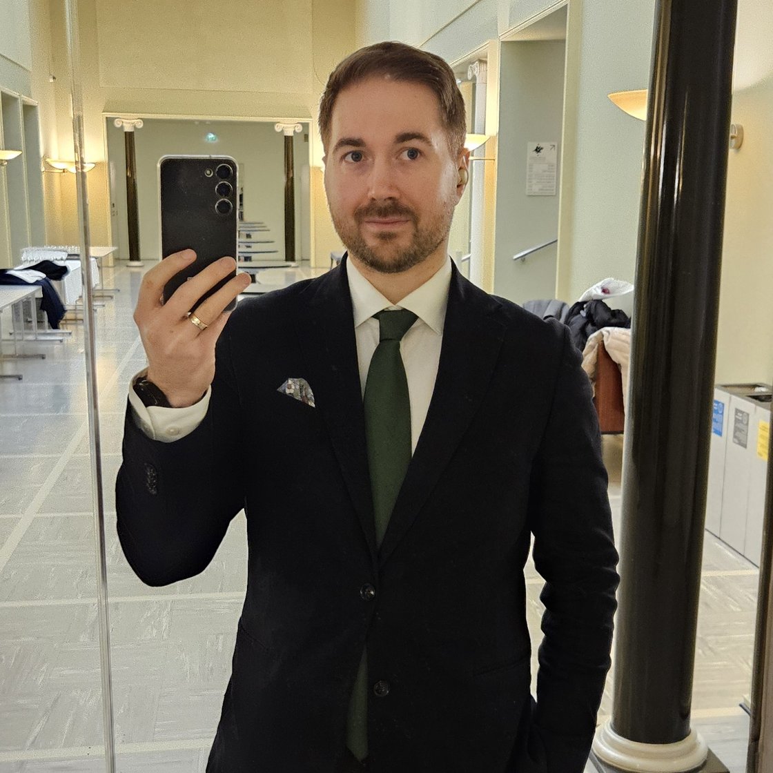 Lauri Lavanti seisomassa peilin edessä puhelin kädessä, ottamassa itsestään kuvaa. Päällä musta takki, valkoinen kauluspaita ja kaulassa vihreä kravatti. Taustalla Helsingin Yliopiston päärakennuksen vaalea käytävä.