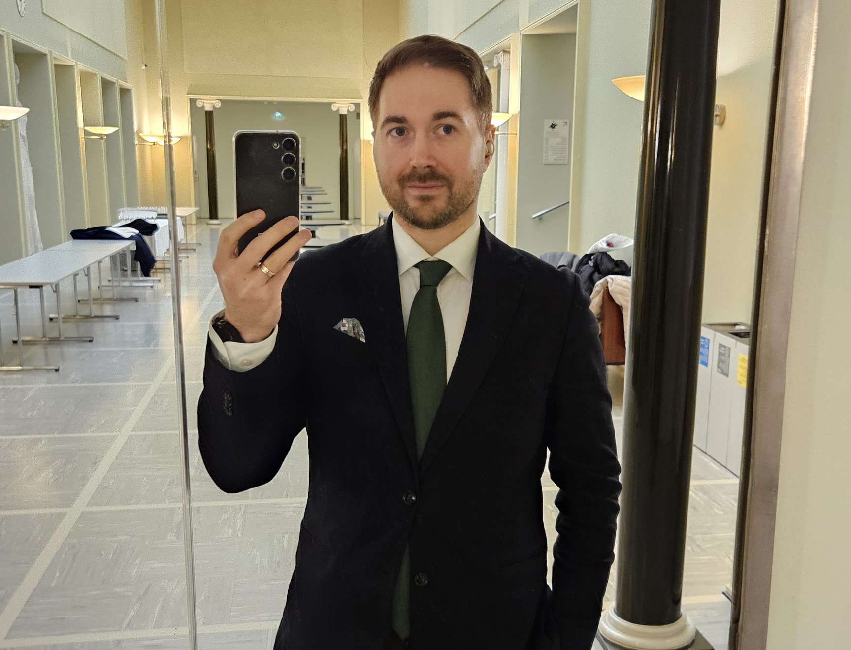 Lauri Lavanti seisomassa peilin edessä puhelin kädessä, ottamassa itsestään kuvaa. Päällä musta takki, valkoinen kauluspaita ja kaulassa vihreä kravatti. Taustalla Helsingin Yliopiston päärakennuksen vaalea käytävä.
