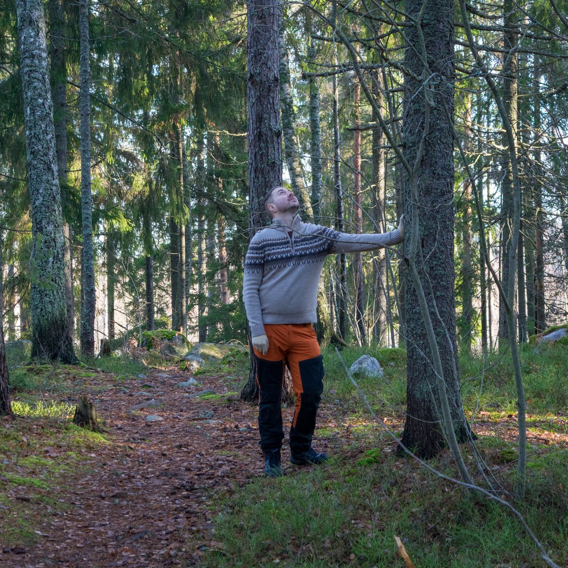 Lauri Lavanti retkeilyvaatteissa metsässä, nojaa puuhun ja katsoo ylös sen runkoa.