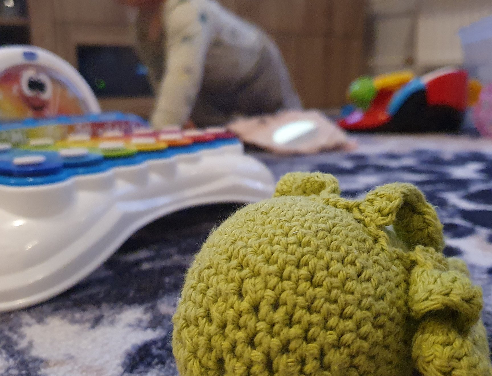 Kuva neulotusta pehmolelusta, taustalla erinäisiä lasten leluja.